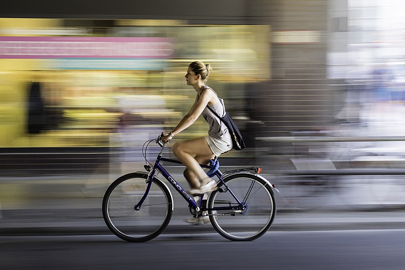 Image is a photo of a woman cycling in an urban street and wearing summer clothes. The photo has been taken as she moves, with the background blurred, giving the photo an added sense of movement. 