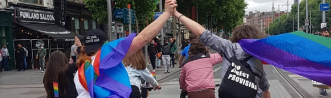 Image is a photo take during last year's Pride Bike Bop by @embeeie. It shows two queer women cycling away from the camera as they travel north along O'Connell Street, Dublin 1. They're wearing pride flags on their backs (like capes, really) and are holding hands in the air. One of the women also has an "End Direct Provision" tote bag on her back, because end Direct Provision, ffs.